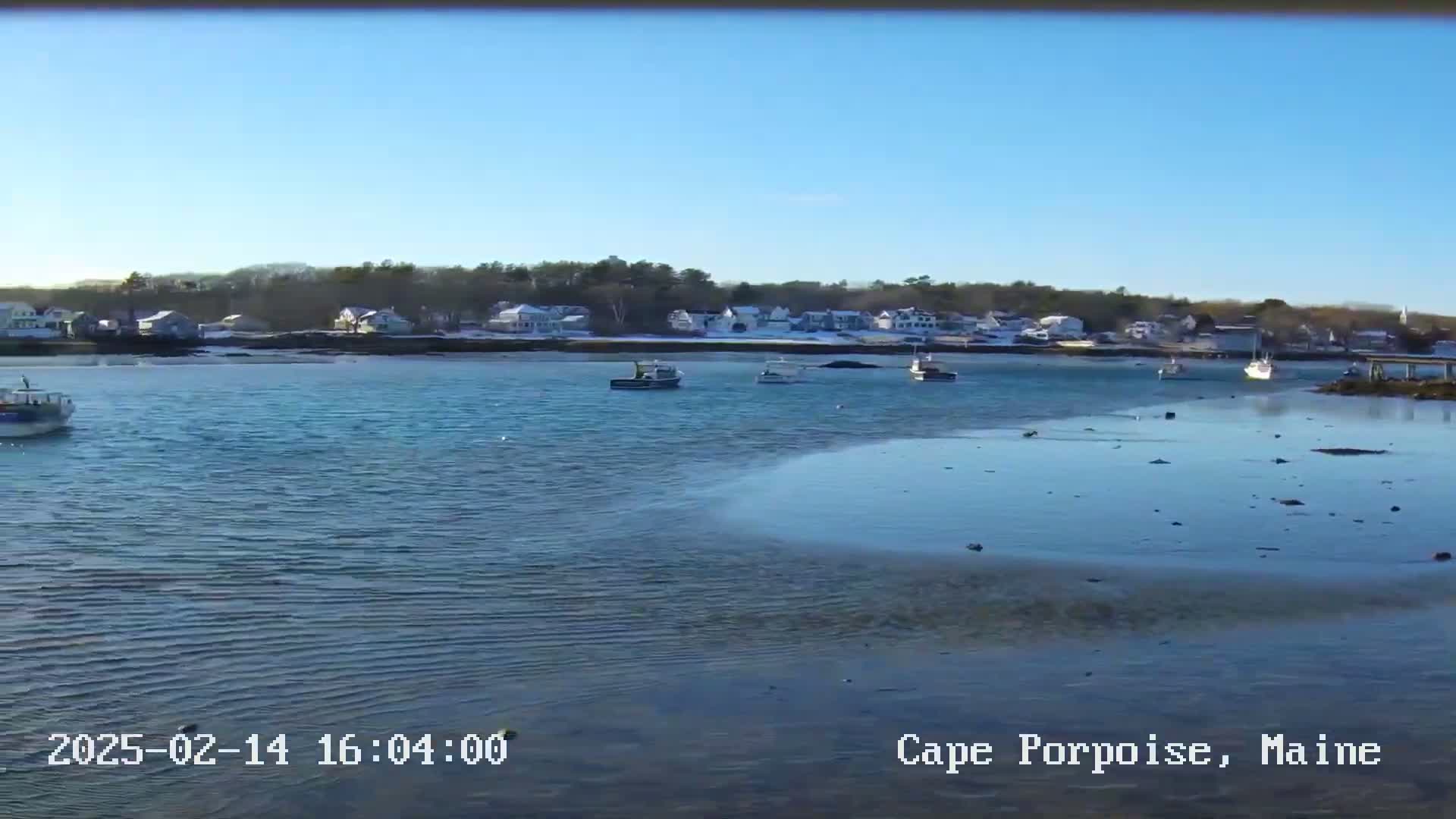 A calm body of water with several small boats and a shoreline of houses with snow-covered ground under a clear blue sky.