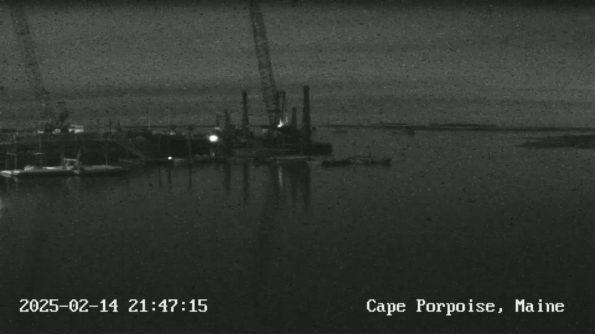A nighttime, low-light image shows a dark body of water with several boats and a construction crane visible near a dock.