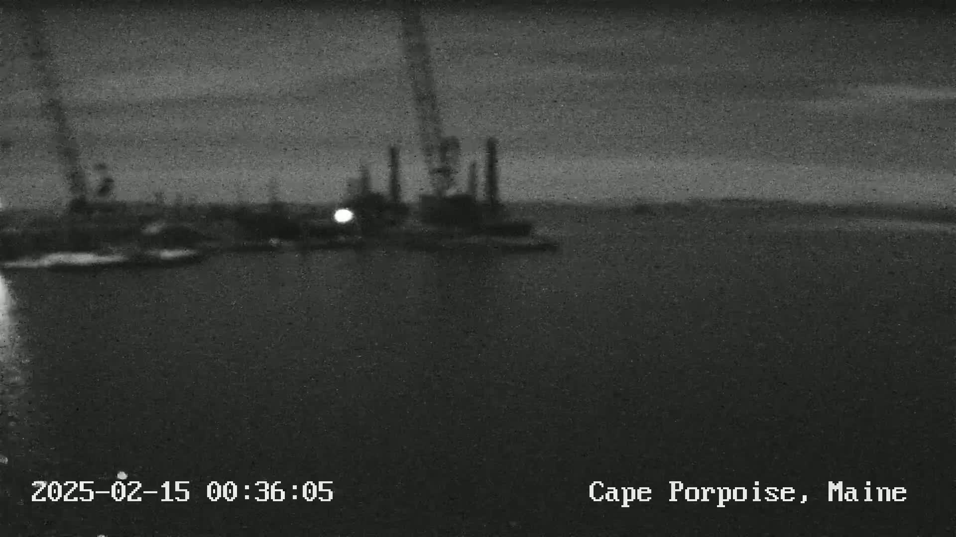A nighttime, low-light image shows an industrial area near a body of water, possibly a harbor, under a dark, overcast sky.