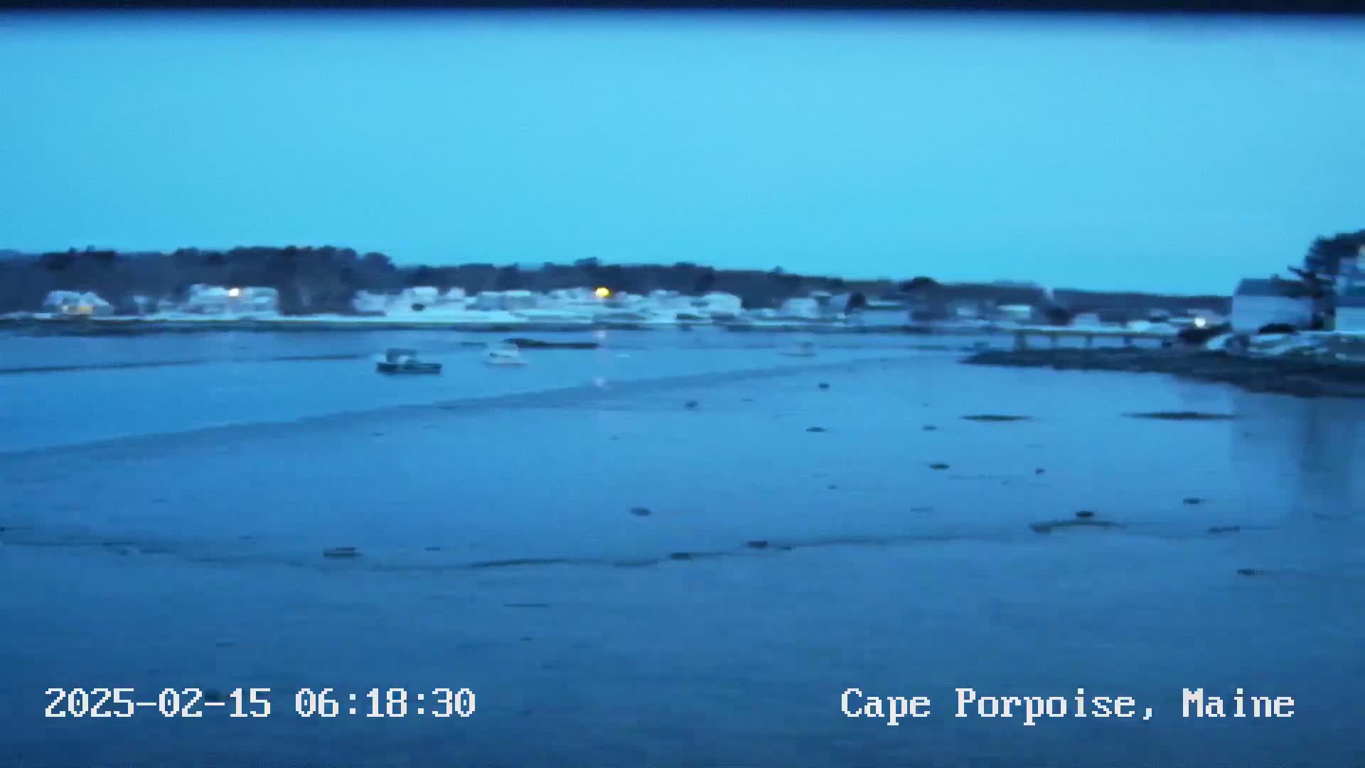 A calm, blue body of water shows a few small boats and a snow-covered shoreline at dusk.