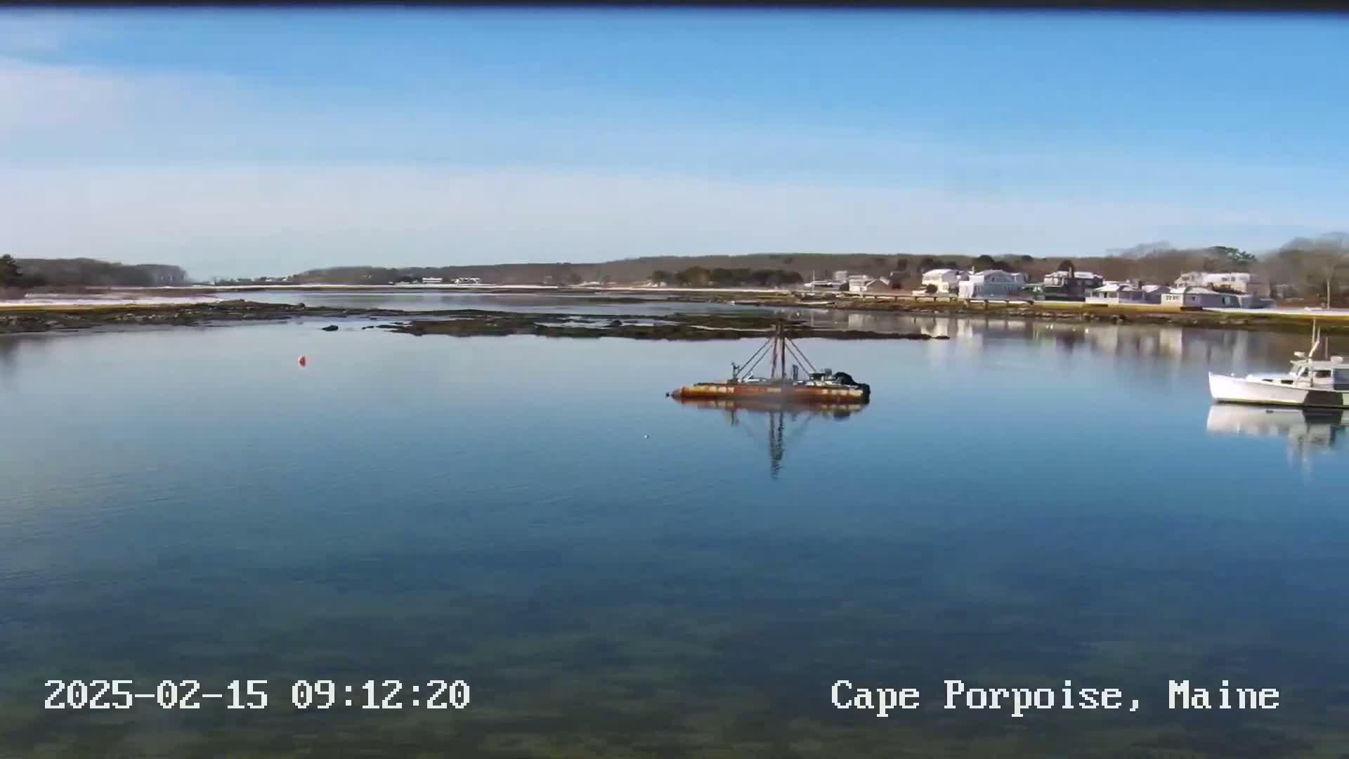 A calm, clear day reveals a waterway with a small barge and a boat, alongside a shoreline featuring houses and a hint of snow on the ground.