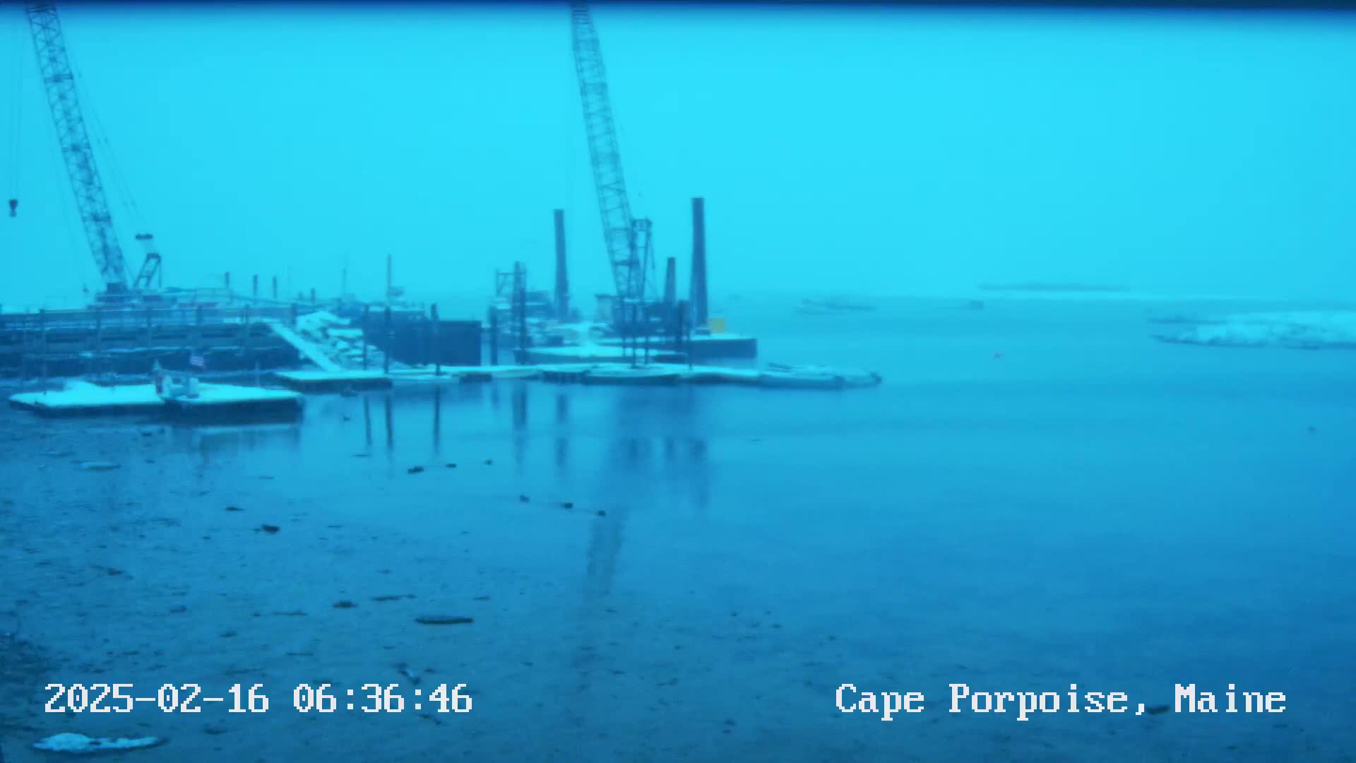 A snow-covered dock with construction cranes and pilings is reflected in calm, blue water under an overcast sky.