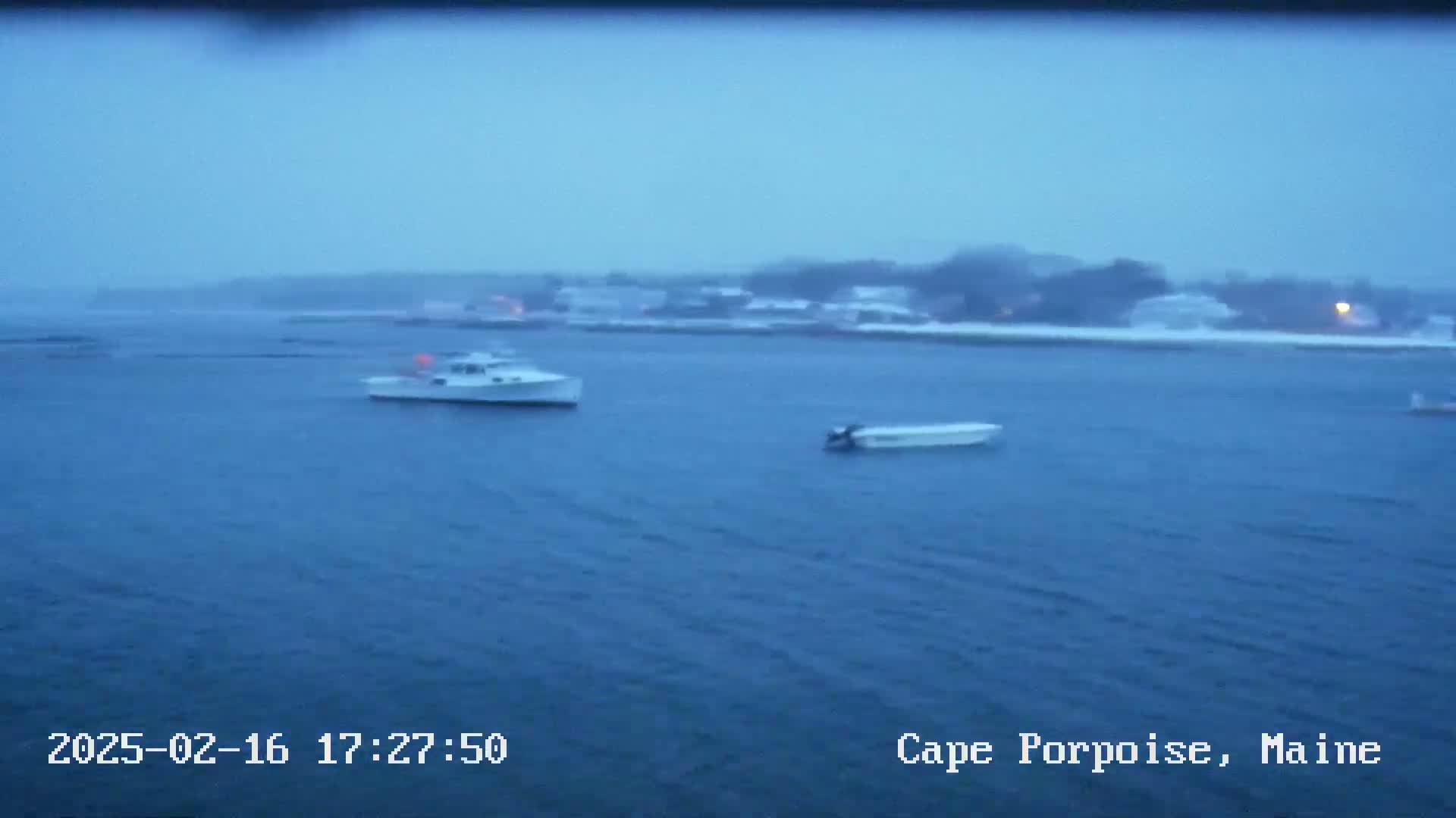 Two small boats are on a body of water in front of a snow-covered shoreline under an overcast sky.