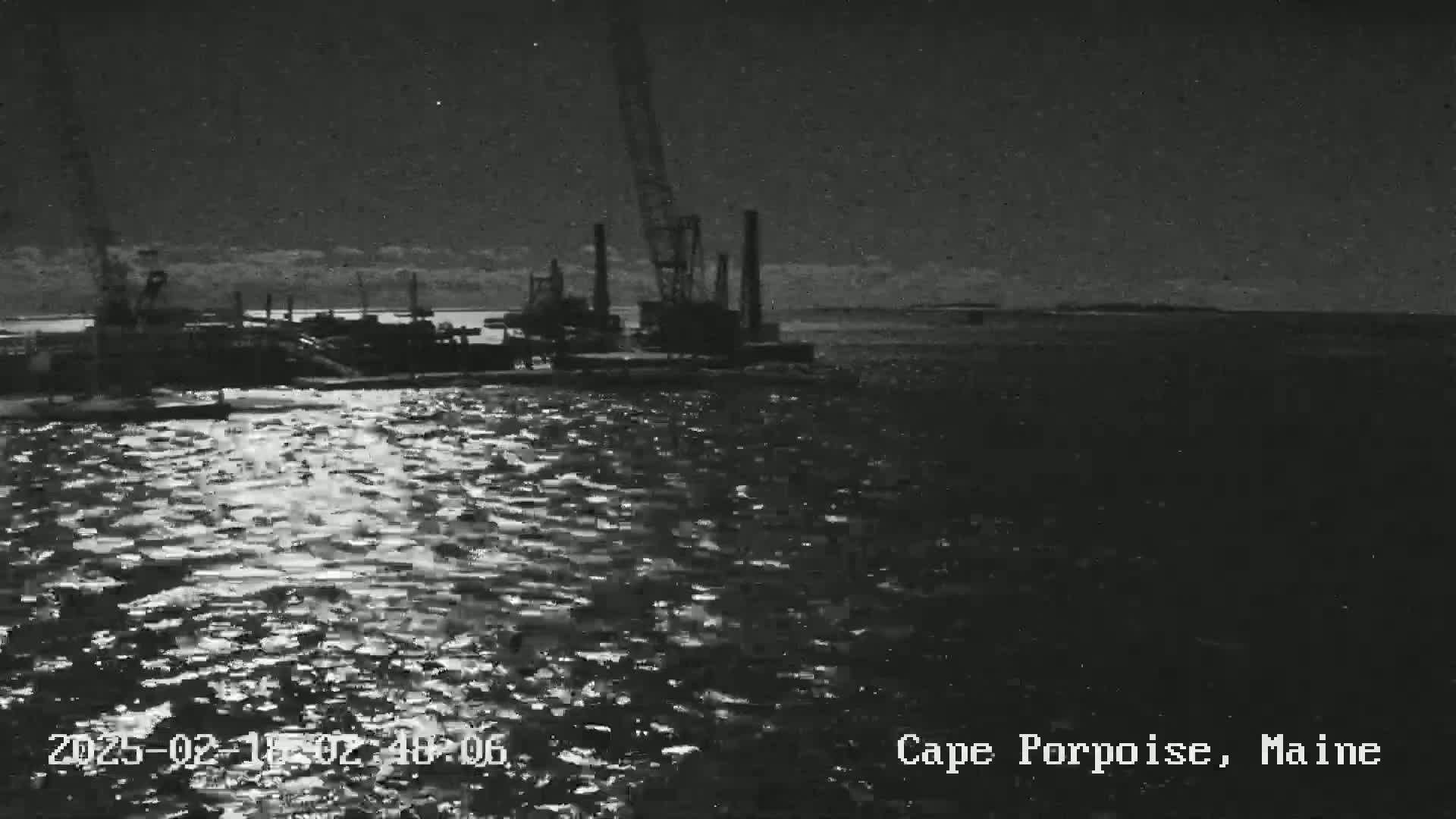 A nighttime, low-light image shows a large piece of industrial equipment, possibly a crane, on a body of water under a mostly cloudy sky.