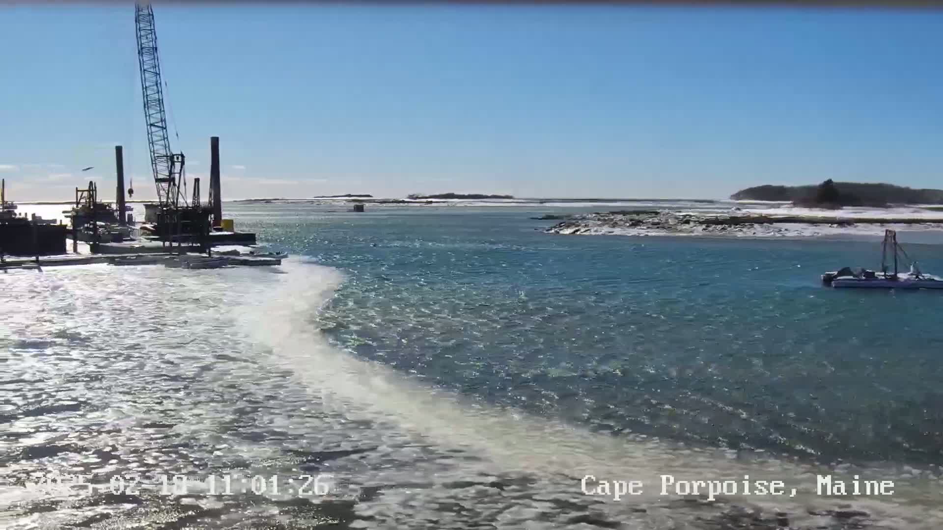 A partially frozen body of water with a construction crane and a small boat visible under a clear, sunny sky.