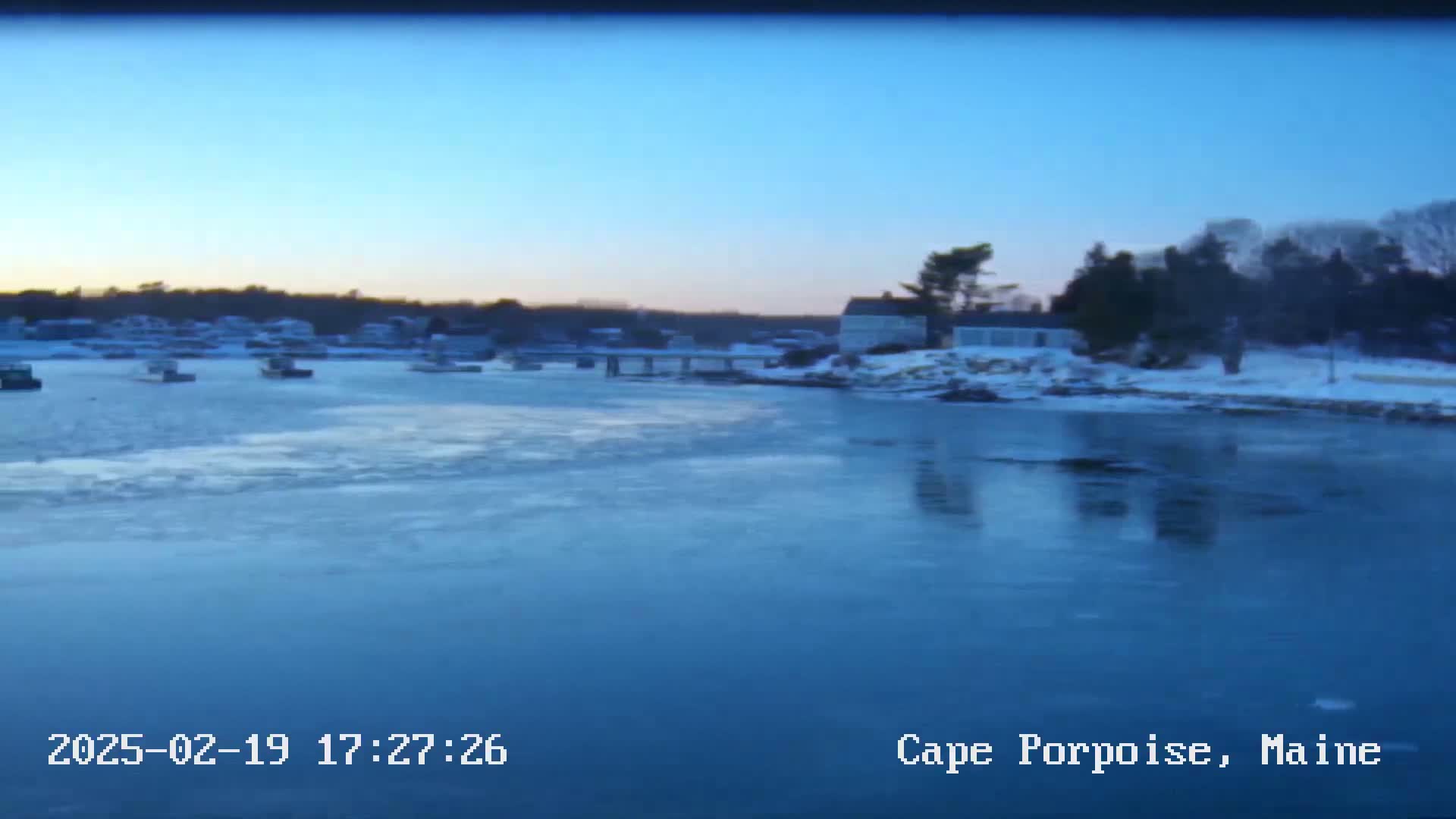 A partially frozen body of water with several boats and a snow-covered shoreline at dusk.