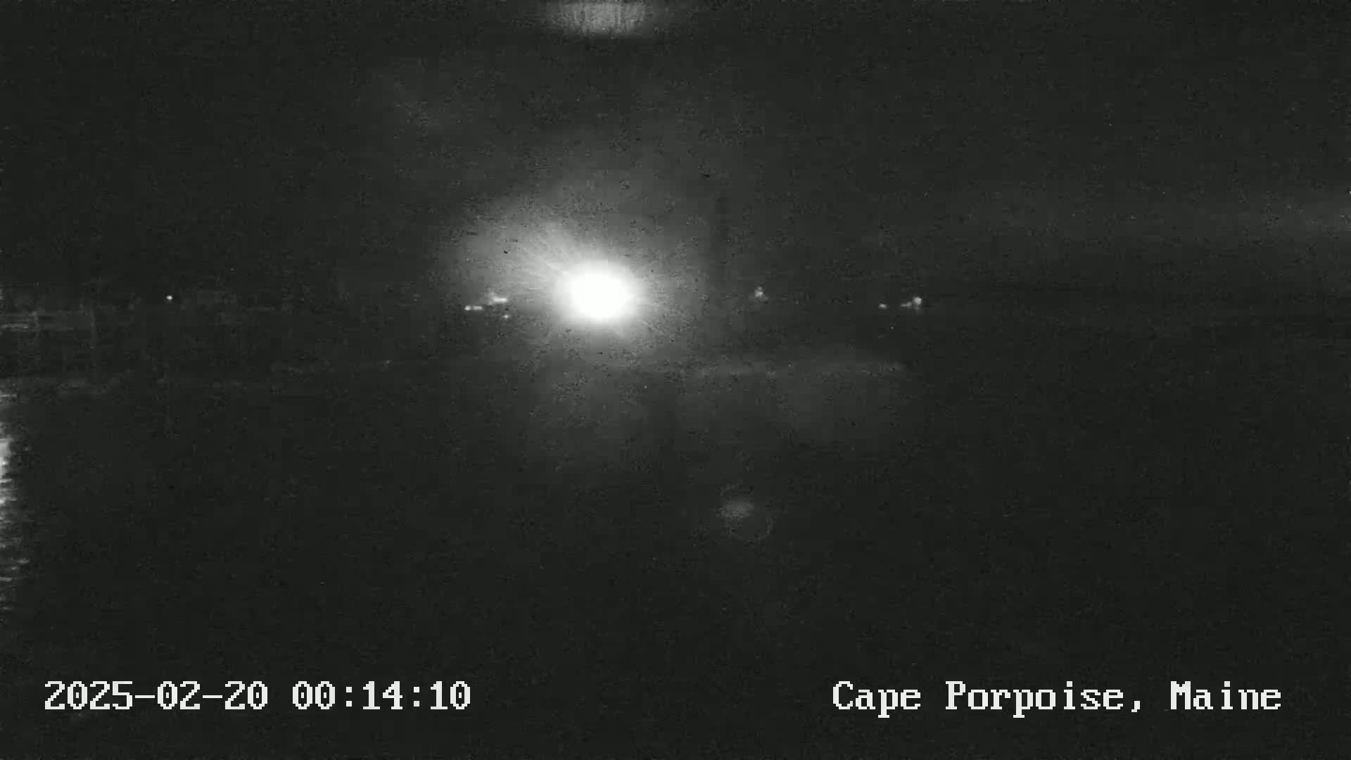 A nighttime, low-light image shows a bright light in the center and faint, dark shapes of buildings or structures along a dark body of water.