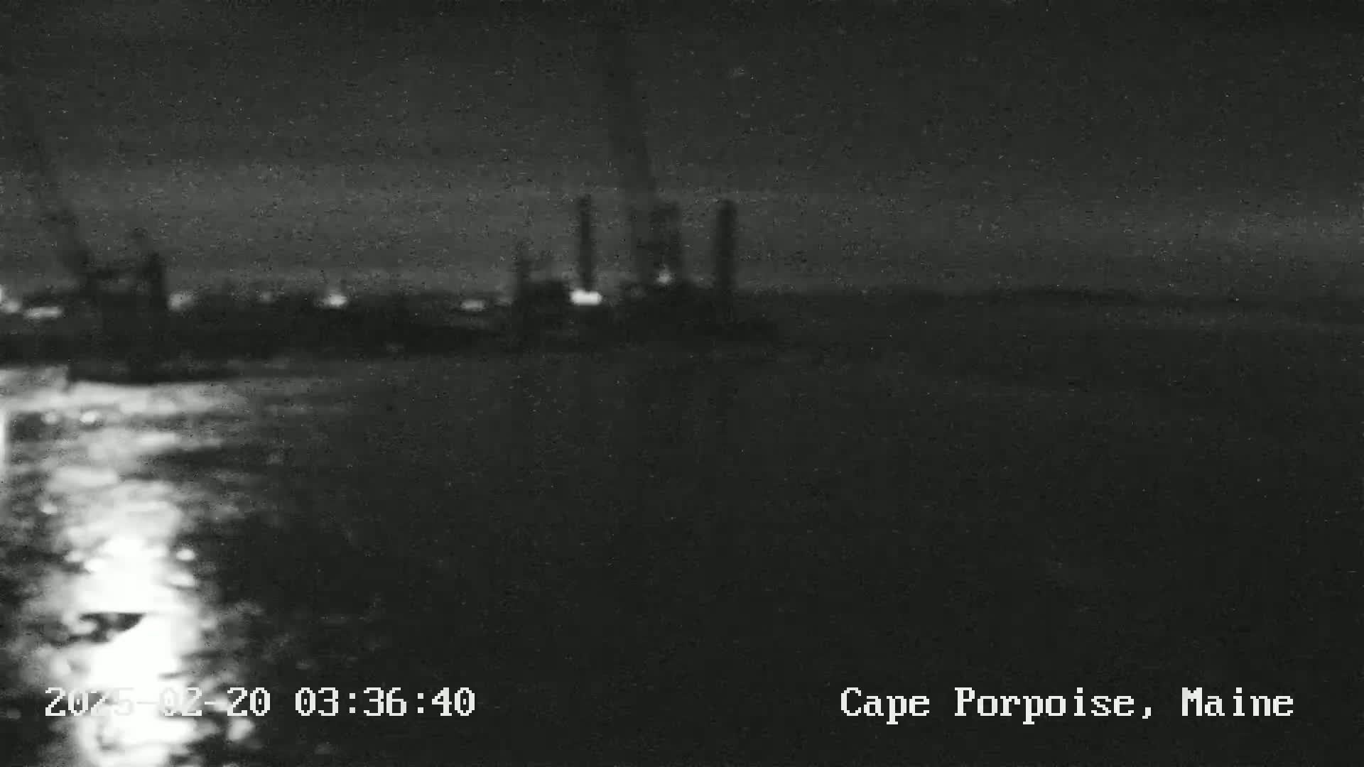 A blurry, nighttime image shows an industrial area across a dark body of water under a mostly clear sky.