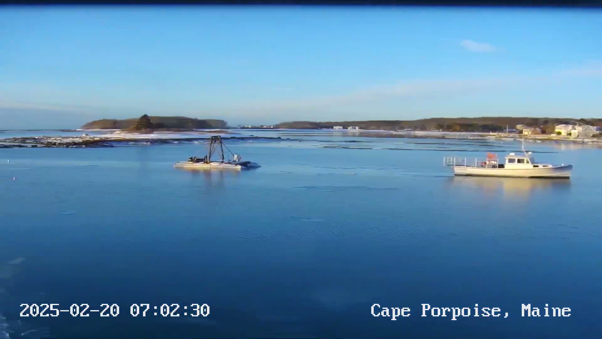 A calm, blue body of water with a small boat and a piece of floating equipment is visible, with a snow-covered shoreline and distant trees under a clear, sunny sky.