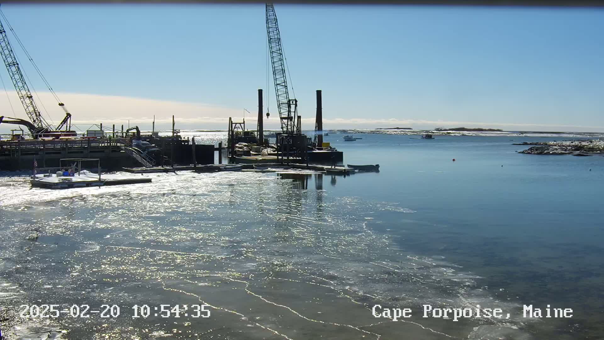 A large crane sits on a barge in a harbor partially covered in ice on a sunny day.