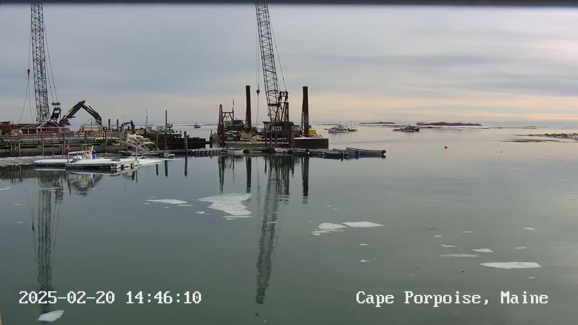 A calm harbor with scattered ice floes reflects construction equipment and several boats under an overcast sky.