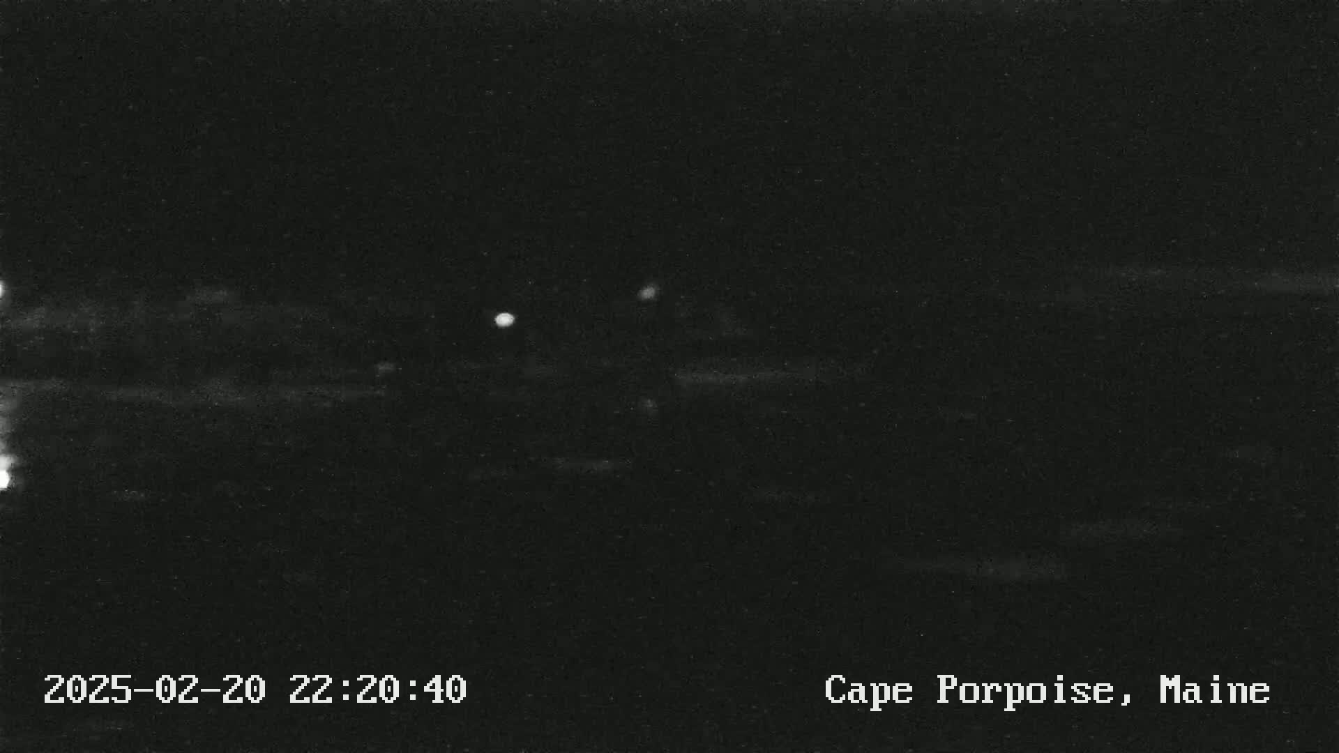 A nighttime, low-light image shows a dark landscape with a few faint lights in the distance.