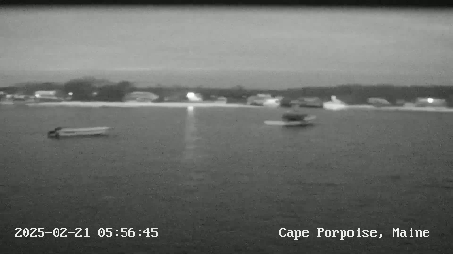 A nighttime, low-light image shows a body of water with a few small boats, and a shoreline with dimly lit buildings in the background.