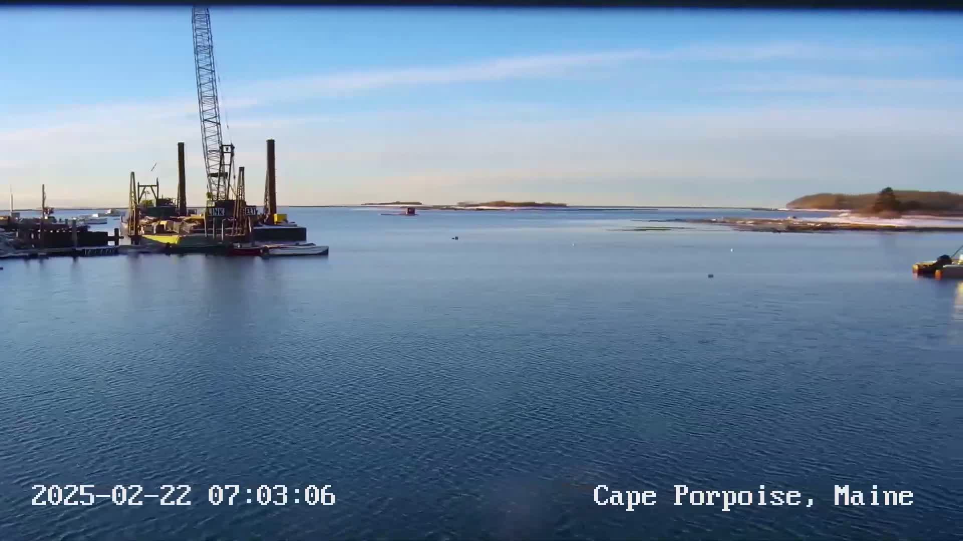A large crane and other construction equipment are situated in a calm body of water under a clear blue sky.