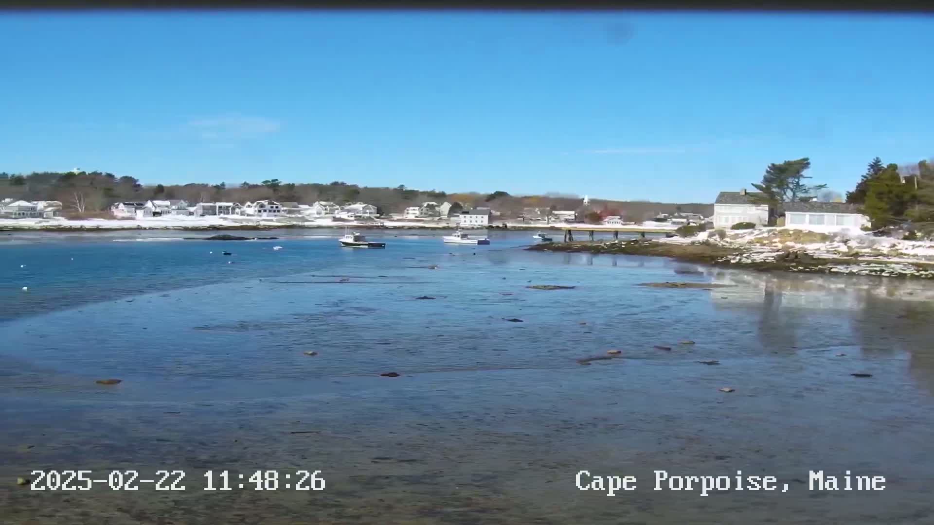 A calm, sunny day reveals a coastal town with a few boats in the harbor, partially covered in snow.