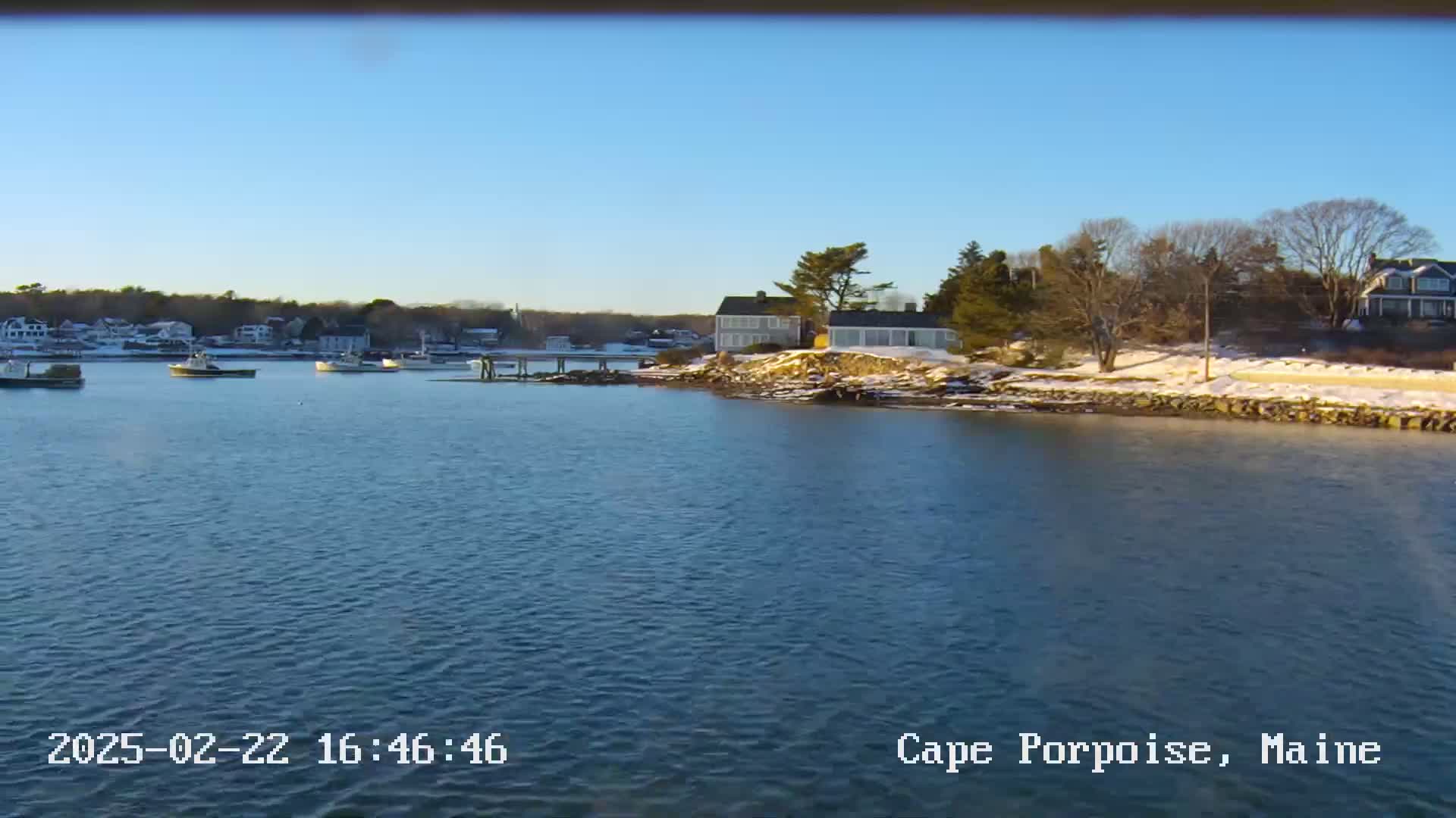 A calm body of water shows several boats near a shoreline with houses and patches of snow under a clear blue sky.