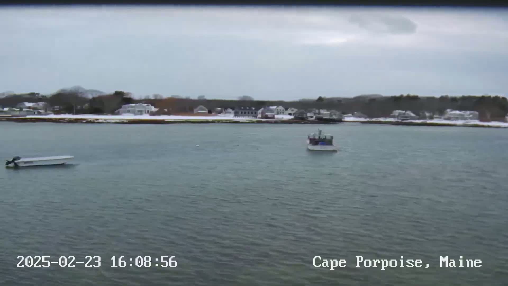 A calm body of water with two small boats, a snowy shoreline with houses and trees visible in the distance under an overcast sky.