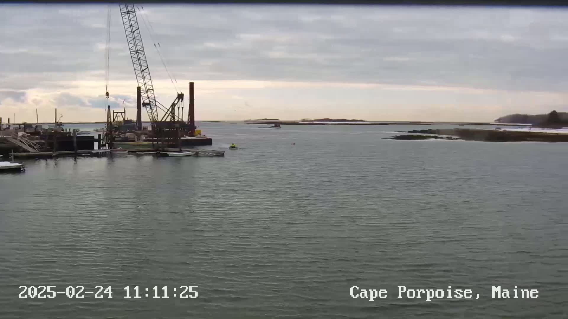 A large crane is positioned near a harbor on a cloudy day, with a calm body of water extending to a distant shoreline.
