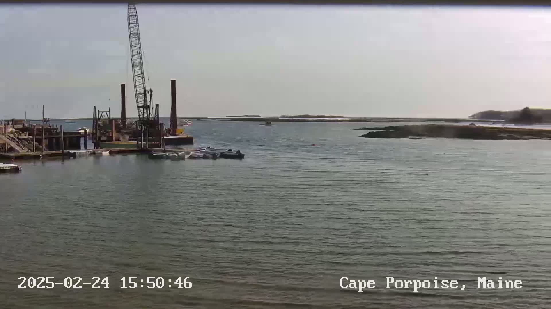 A harbor scene on a calm, overcast day shows a large crane and several boats near a dock with rocky land and distant coastline in the background.