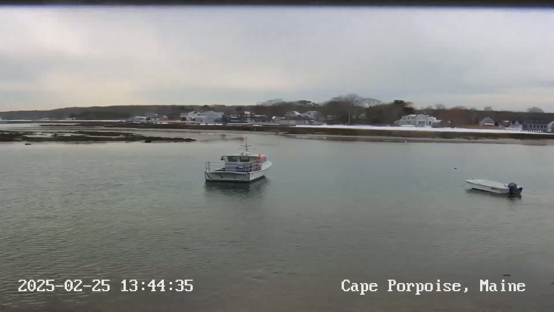 A calm body of water with a larger boat and a smaller boat, under an overcast sky with a snow-covered shoreline in the background.
