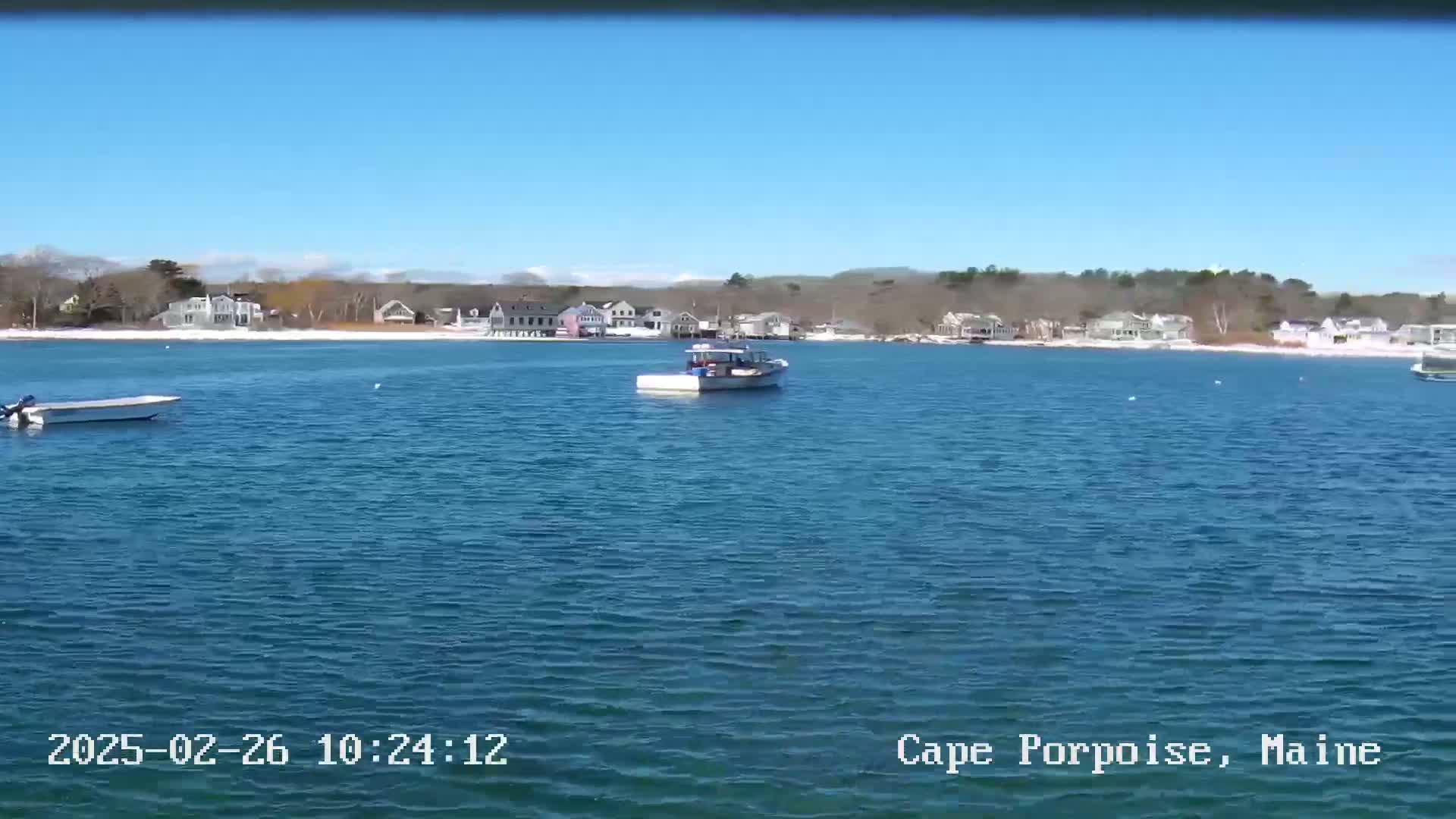A calm, blue body of water with a few small boats and a shoreline of houses and trees under a clear blue sky.