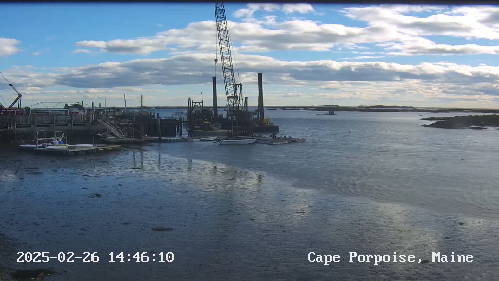 A construction crane and several small boats are situated at a harbor under a partly cloudy sky.