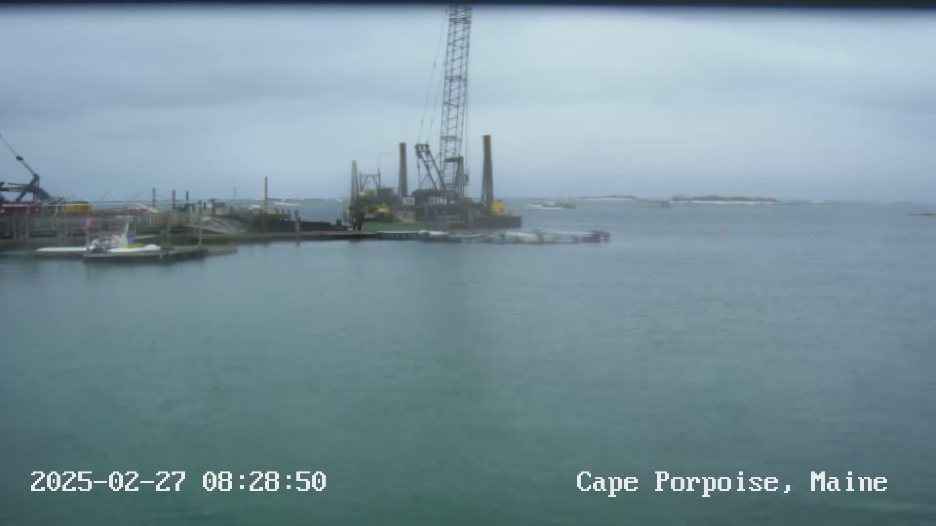 A large crane sits on a barge in a harbor on an overcast day, with several smaller boats moored nearby.