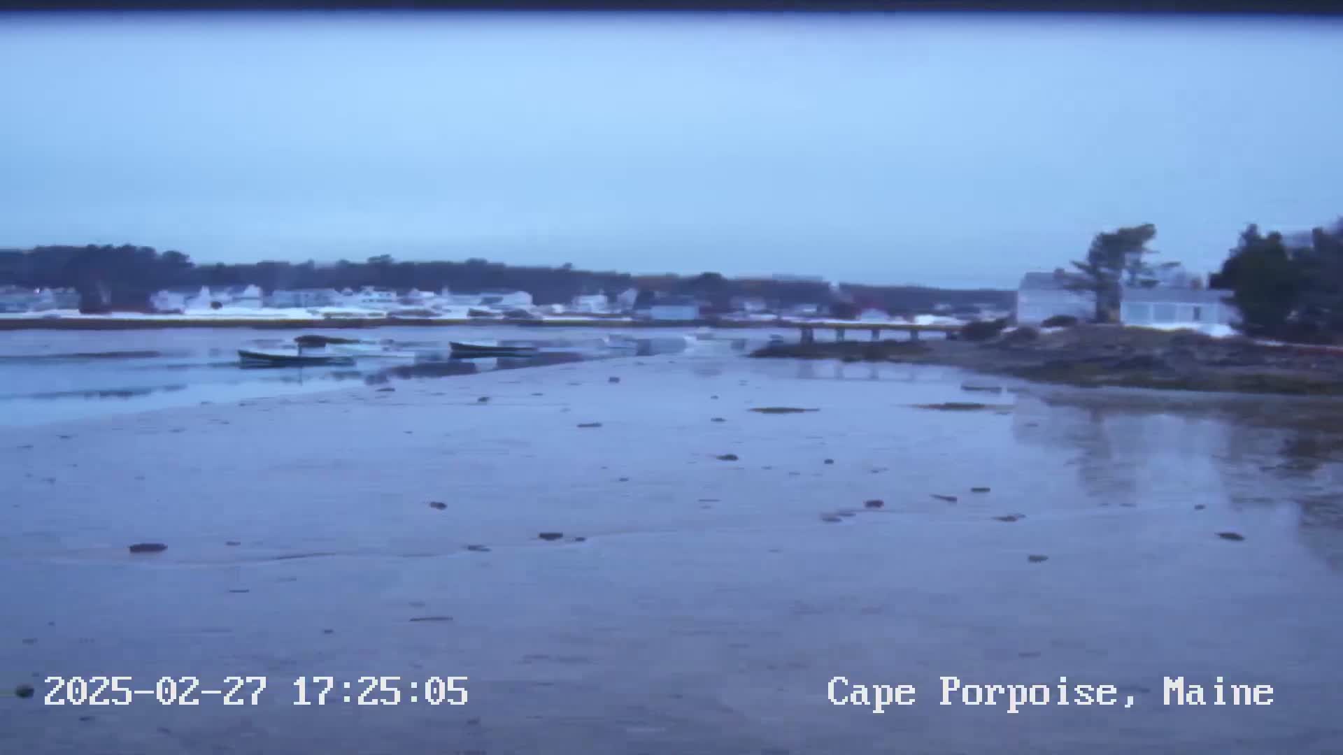 A calm, overcast day reveals a tidal flat with several small boats moored near a shoreline with houses and trees.