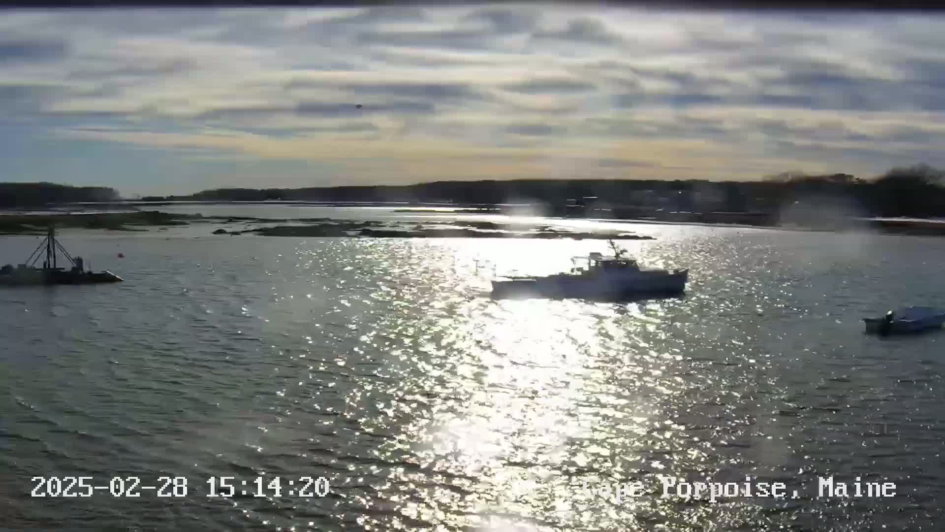 A partly sunny day shows three boats on calm, shimmering water near a shoreline with trees.