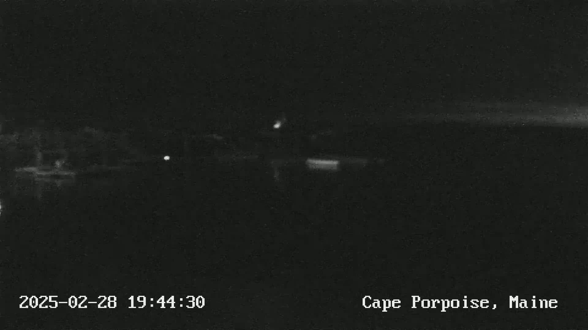 A dark, nighttime scene shows a body of water reflecting a few faint lights, with a dark shoreline in the background.