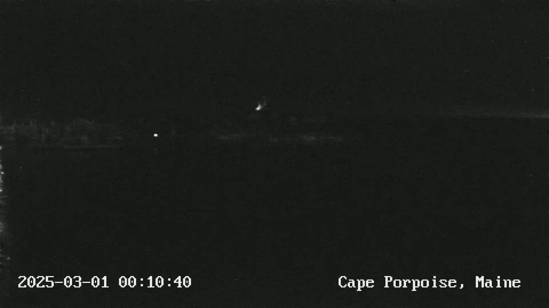 A nighttime, low-light image shows a dark landscape with a few faint lights visible in the distance.