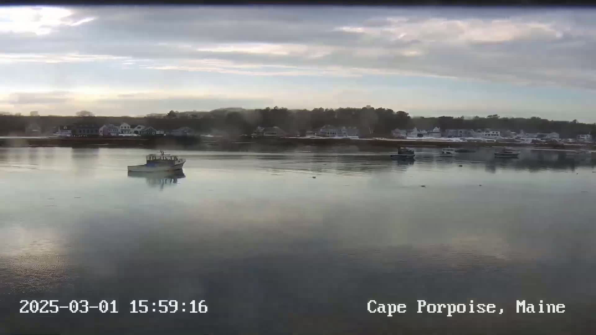 A calm body of water shows several small boats, with a small town on the far shore under a mostly cloudy sky.