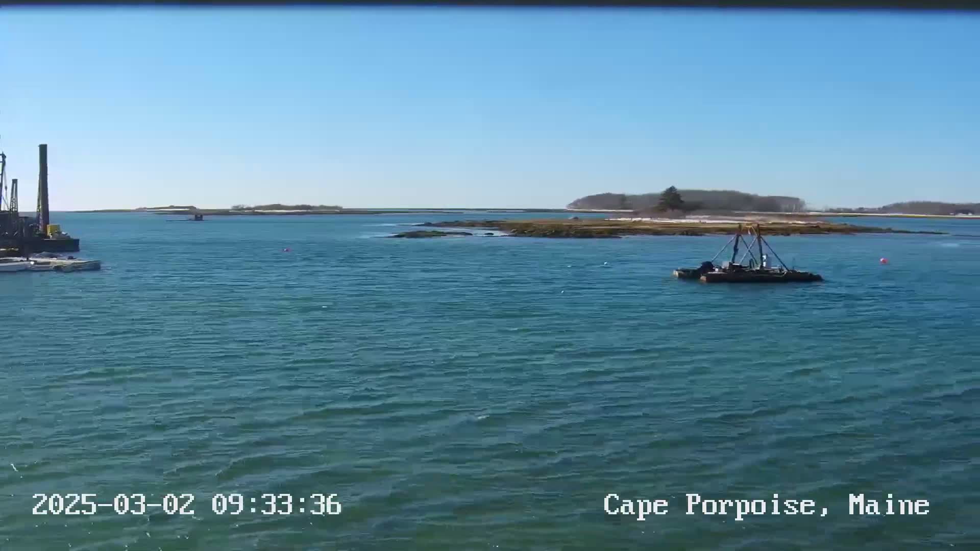A calm, sunny day shows a body of water with two small work boats and a small island in the distance.
