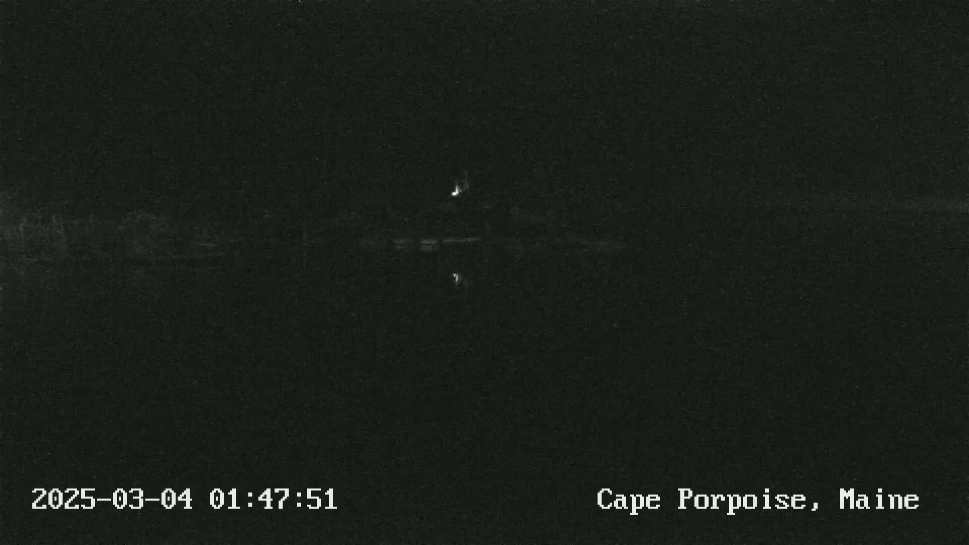 A dark, nighttime image shows a body of water reflecting a single light source on the opposite shore.