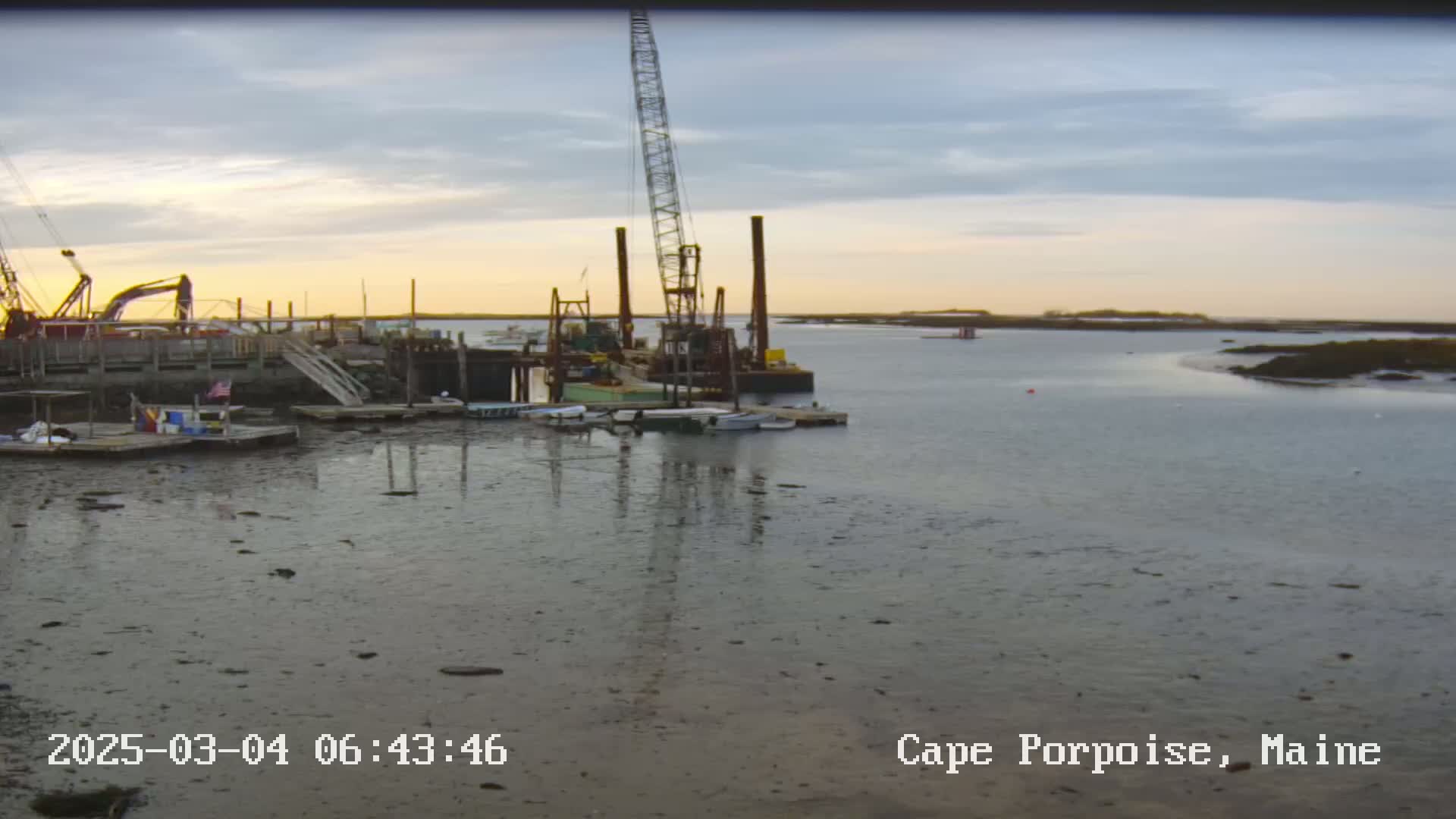 A calm, dusky-lit harbor scene shows a construction crane and several boats moored at a dock, with exposed mudflats at low tide.