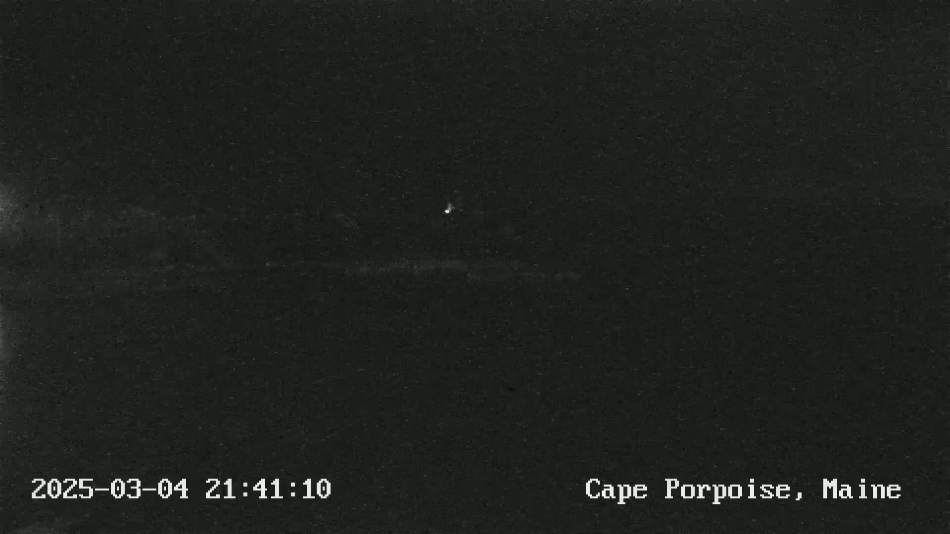 A nighttime, low-light image shows a dark landscape with a single, faint light source in the distance.