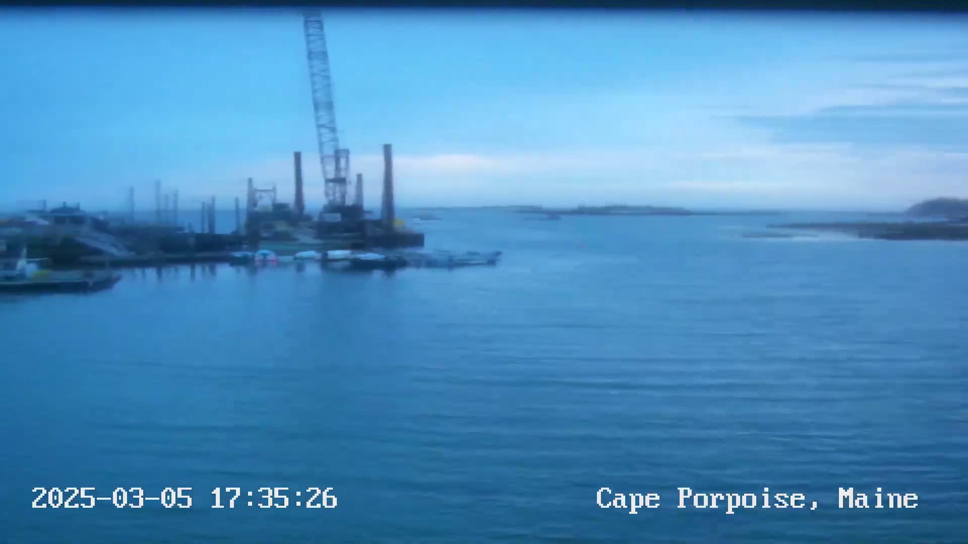 A mostly calm body of water shows a crane and several small boats near a shoreline under a partly cloudy sky.