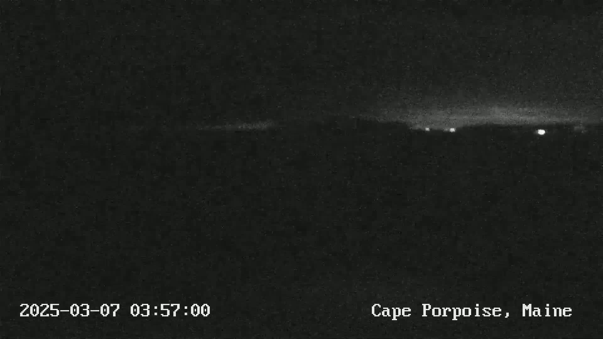 A nighttime, low-light image shows a dark landscape with a few distant lights under a mostly clear sky.