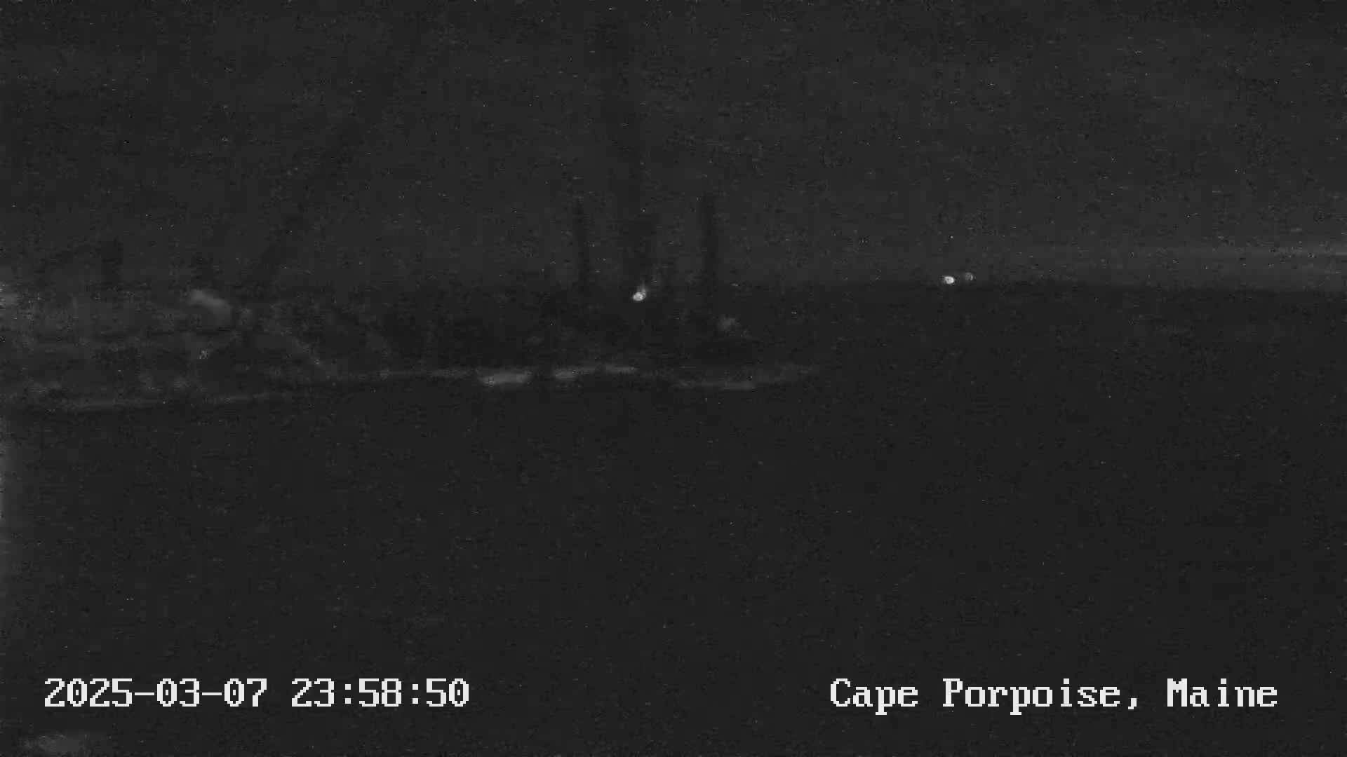 A nighttime, low-light image shows a dark body of water with faintly illuminated structures in the distance.
