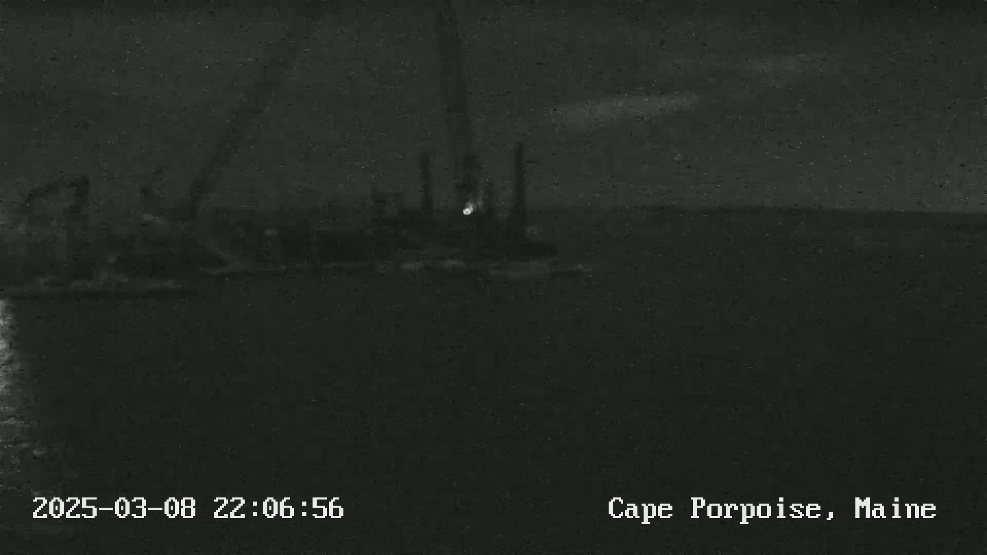 A nighttime, low-light image shows a dark body of water with dimly lit structures or vessels in the distance.