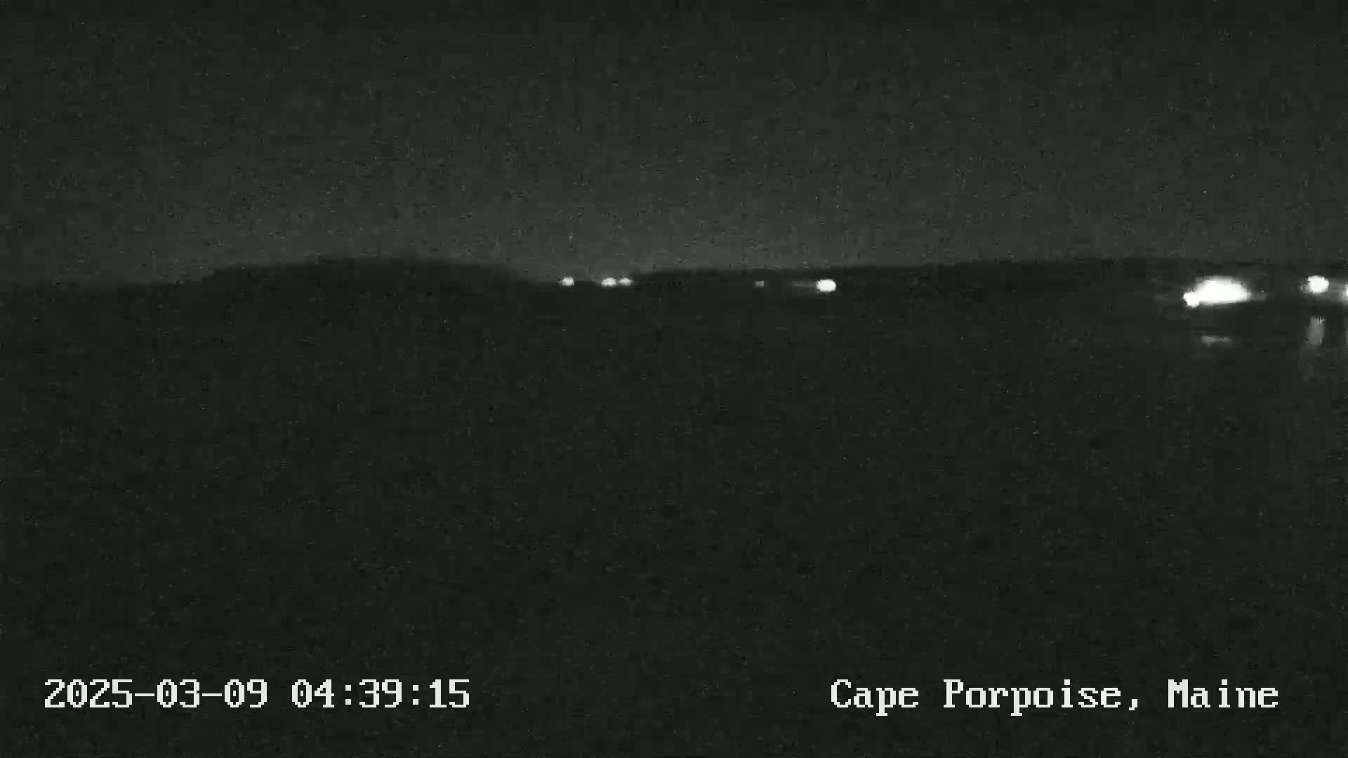 A dark, nighttime landscape shows a distant line of faint lights under a clear, dark sky.