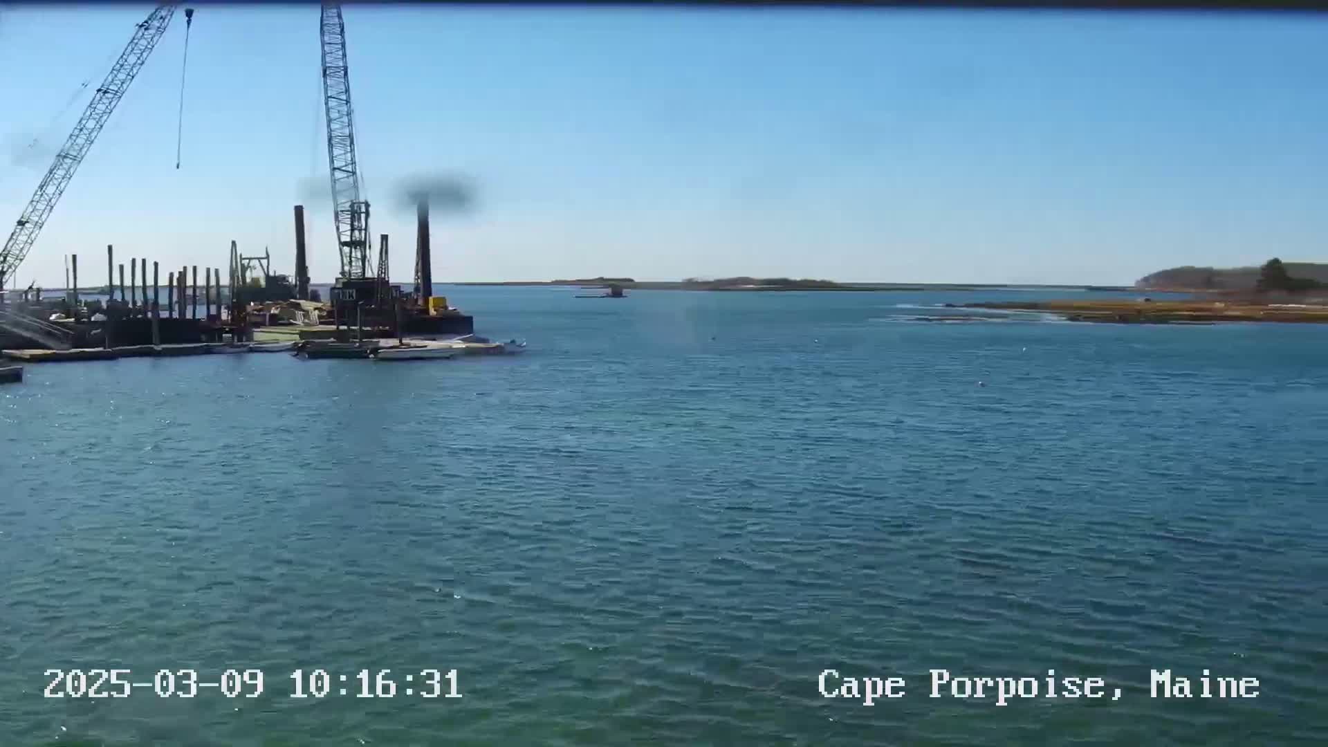 A body of water shows construction equipment and a barge near a shoreline under a clear, sunny sky.