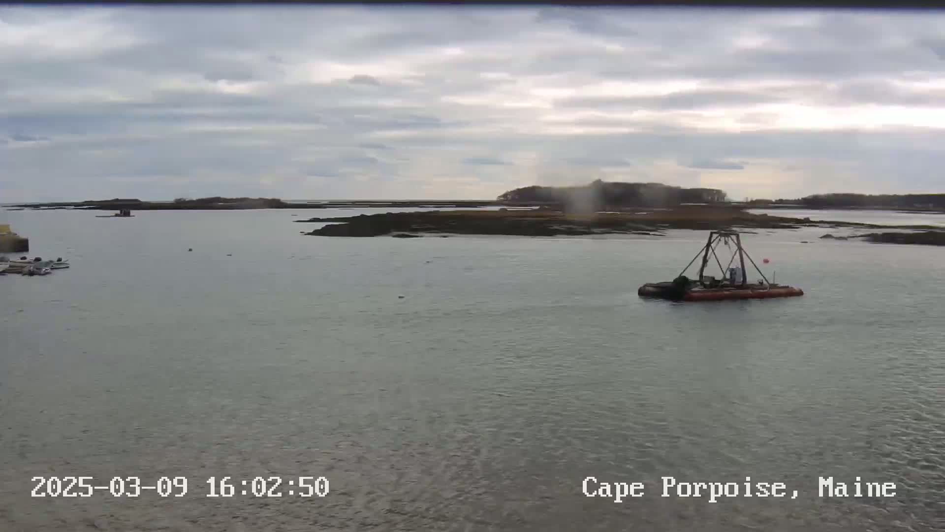 A small boat with a crane is in calm, gray water near rocky islands under an overcast sky.