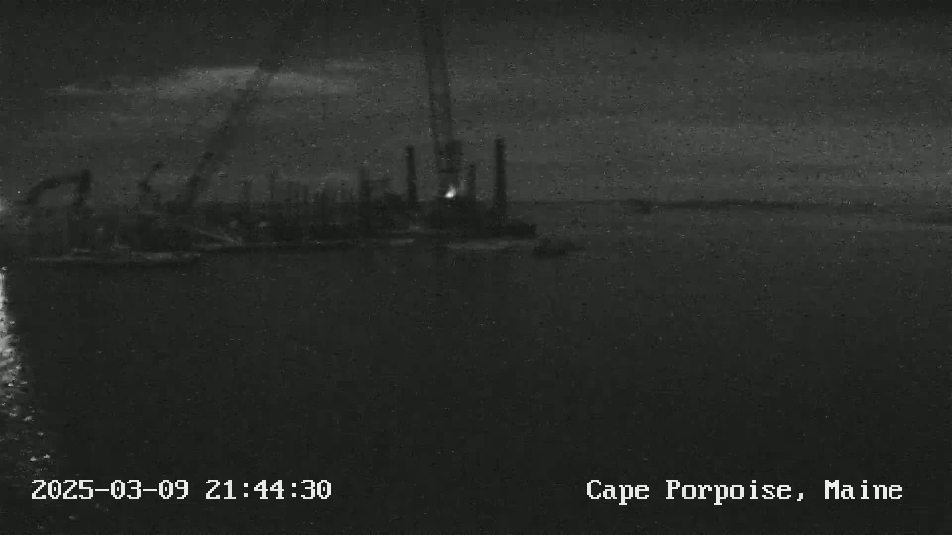 A nighttime, low-light image shows an industrial area with cranes and tall structures near a body of dark water under a cloudy sky.