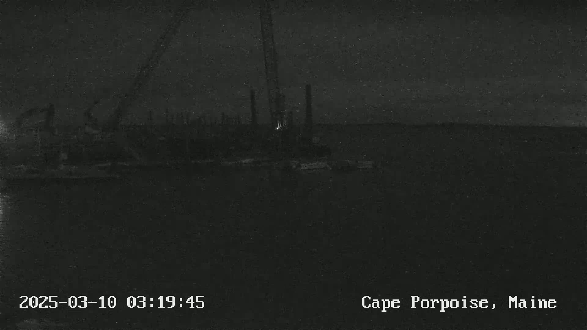 A nighttime, low-light image shows a dark body of water with some indistinct objects and structures near the shore, possibly a construction site.