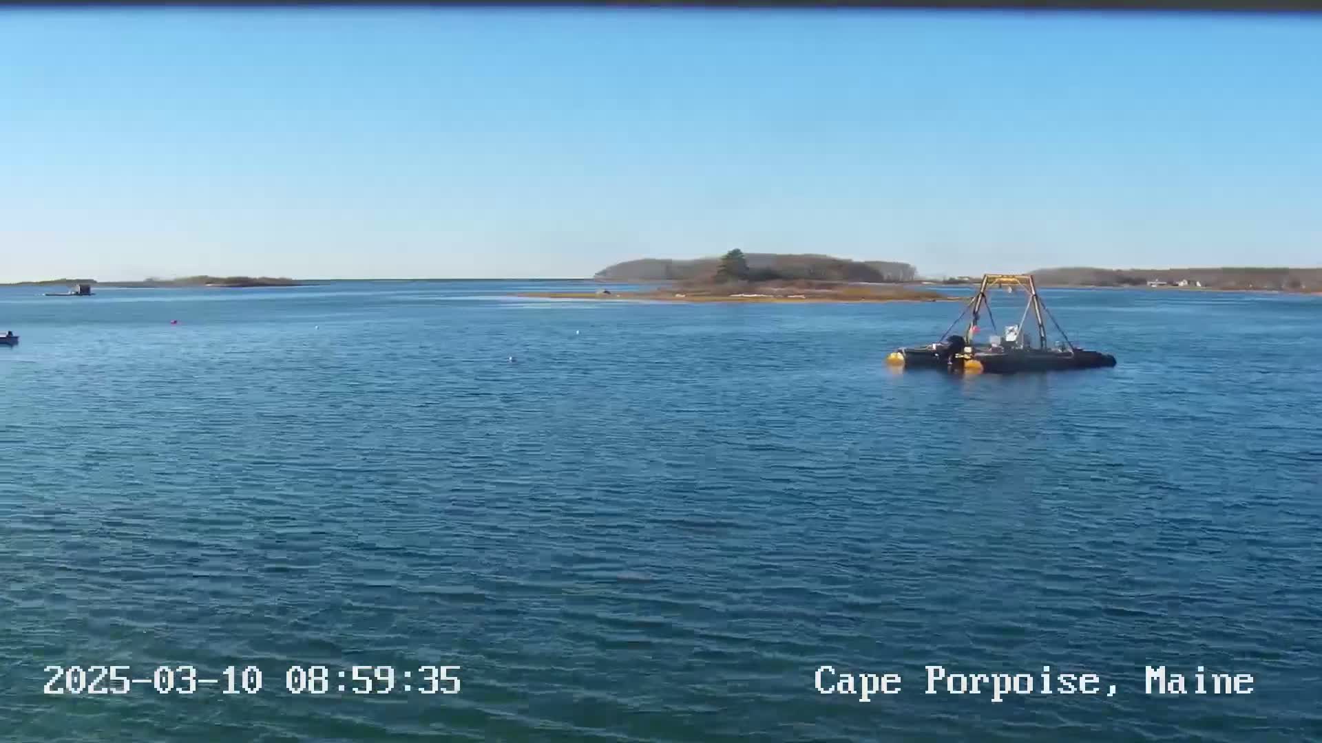 A body of water under a clear blue sky shows a small boat with a crane-like structure in the middle distance, with a small boat and land visible in the far distance.
