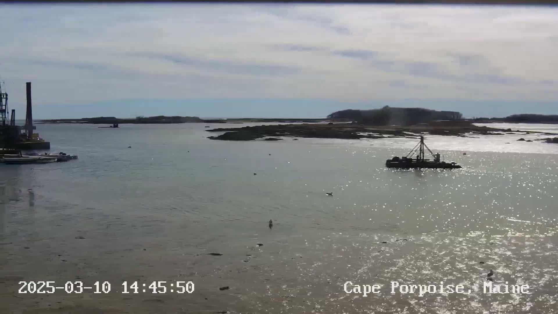 A calm, sunny day shows a body of water with two work boats and a rocky shoreline in the background.