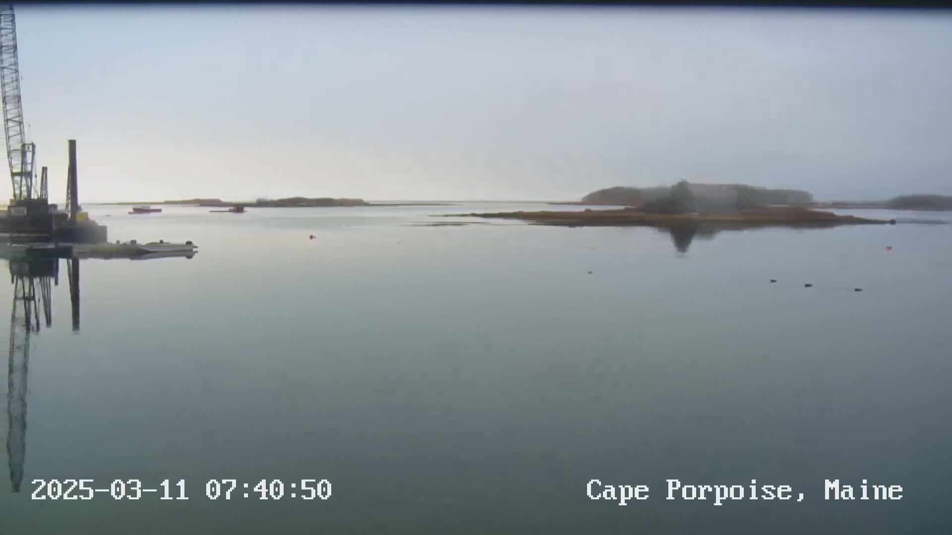 A large crane is situated on the left side of a calm body of water, with several small islands visible in the hazy distance under an overcast sky.