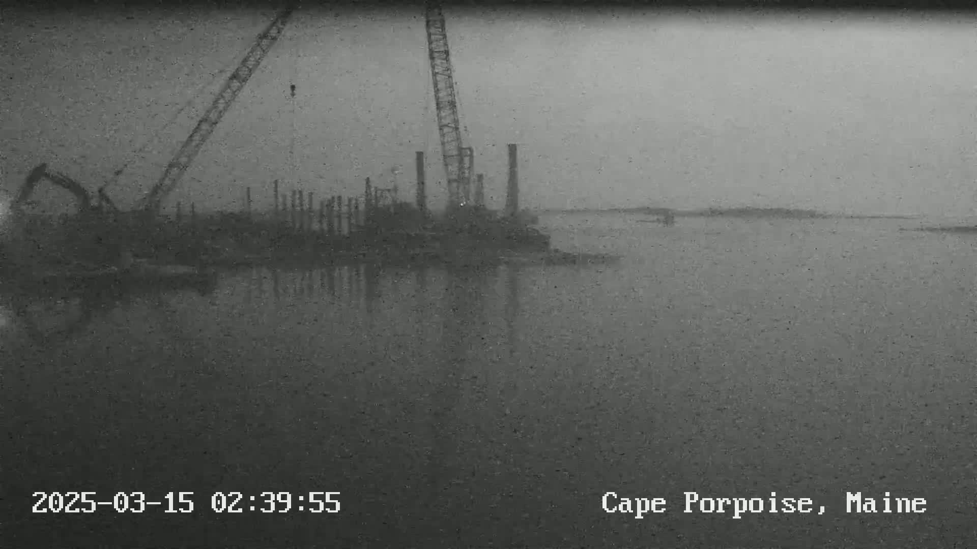 A construction site with cranes and pilings is visible on a calm, gray, overcast day next to a body of water.