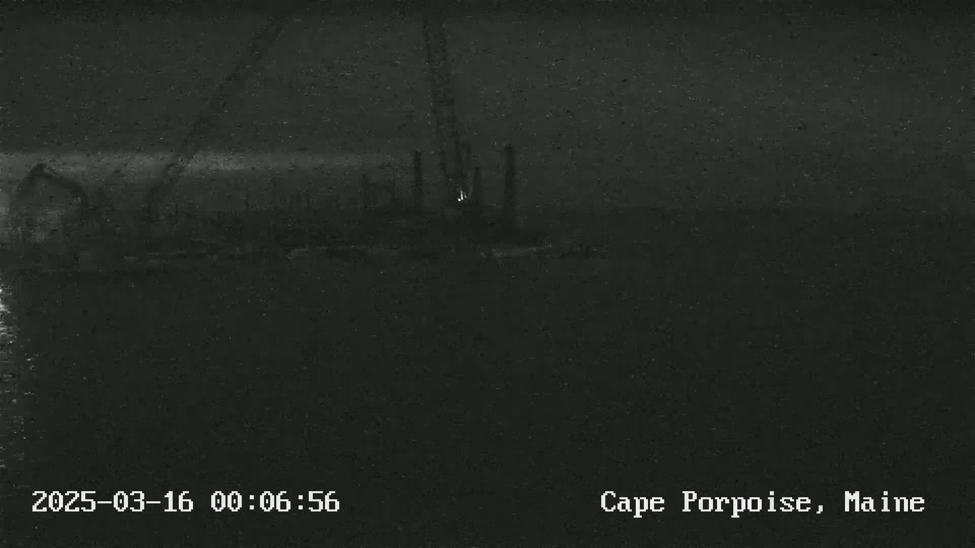 A dark, nighttime image shows an indistinct industrial area near water under a cloudy sky.